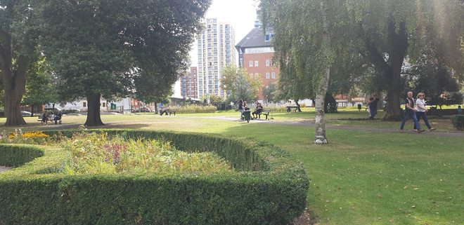 A sunny view across the park's clipped hedges and lawns with trees and tower blocks behind