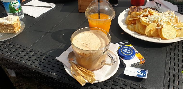 Nachos, a latte in a glass mug, orange juice and a slice of cake on an outdoor table